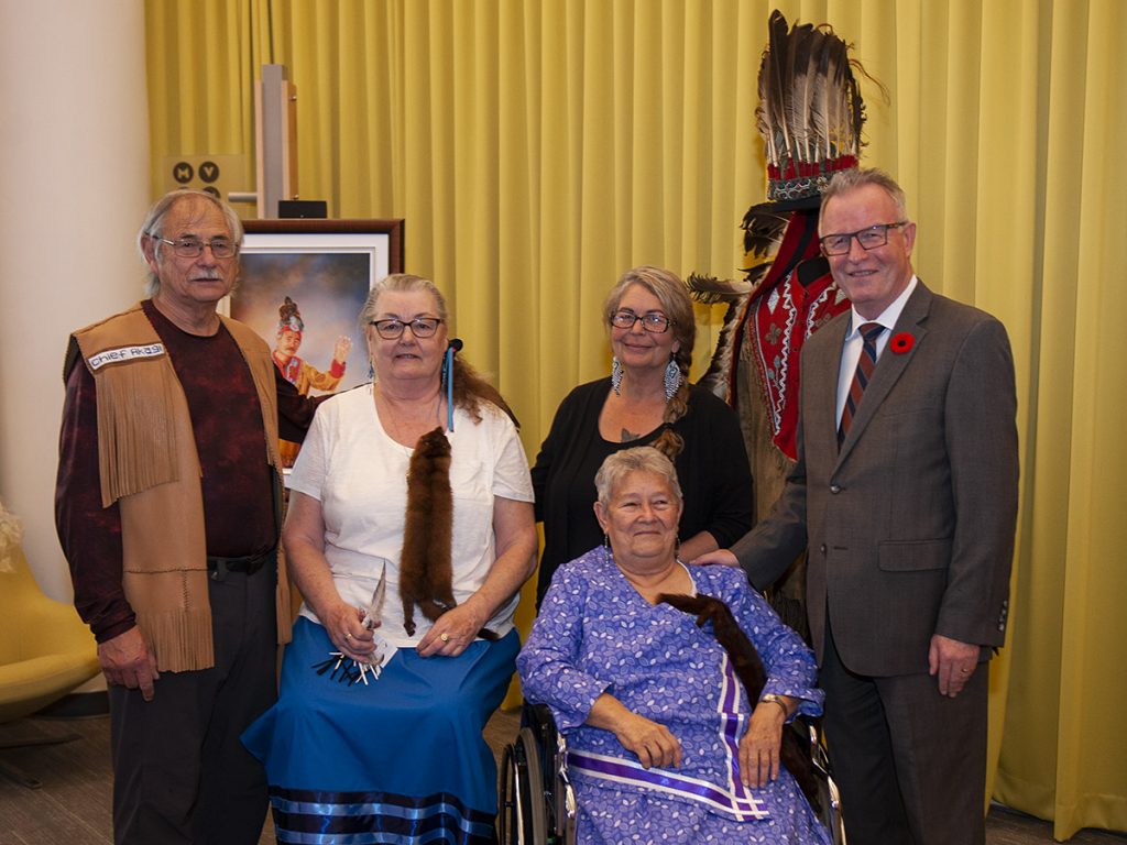 Group of five people, three standing and two seated, including one woman in a wheelchair, in front of a framed image and Indigenous regalia on a mannequin
