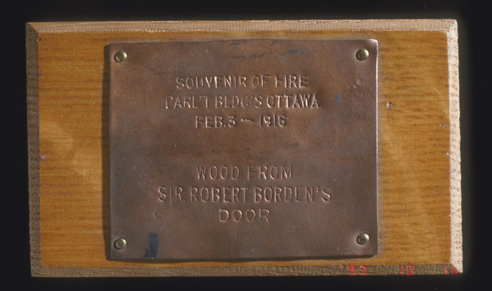 Morceau de bois provenant de la porte du bureau de sir Robert Borden dans la première Chambre des communes. La petite plaque de cuivre porte la mention suivante : « SOUVENIR OF FIRE PARL'T BLDG'S OTTAWA FEB. 3 – 1916. WOOD FROM SIR ROBERT BORDEN'S DOOR. » (Souvenir de l’incendie des édifices du Parlement survenu le 3 février 2016 à Ottawa – bois de la porte du bureau de sir Robert Borden), Musée canadien de l'histoire, 7255-3242-2567-057