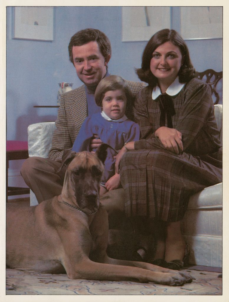 Photograph of 2 adults and a child formally dressed, sitting on a couch along with a great dane.