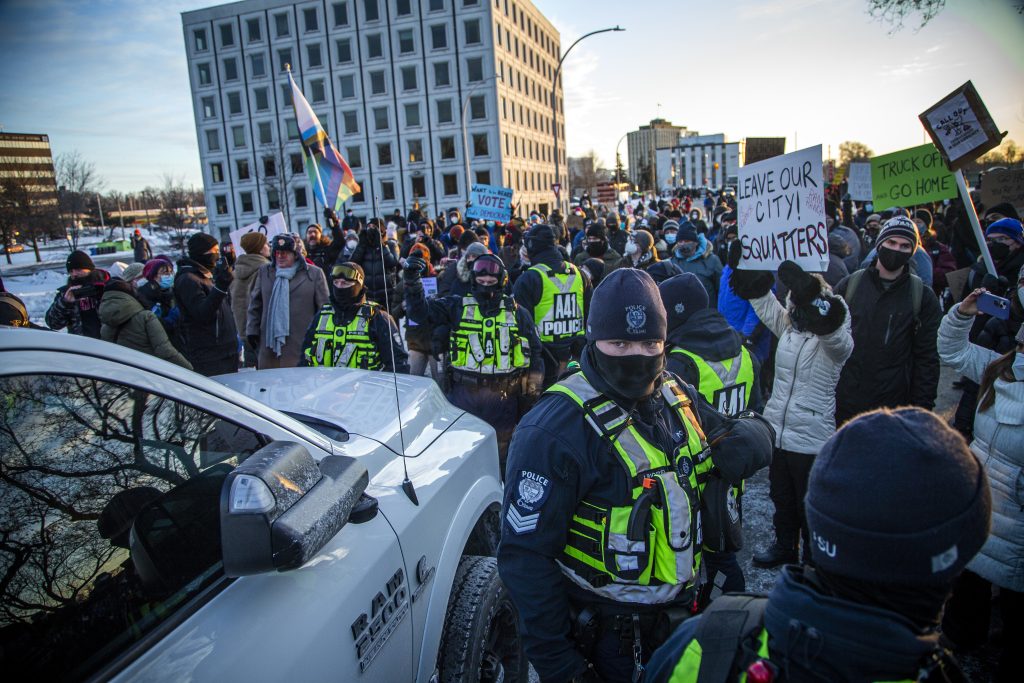 A group of people wearing face masks and winter clothing gather outside, blocking the path of a pickup truck. Placards visible in the crowd read “Leave our city, squatters” and “Truck off go home.”