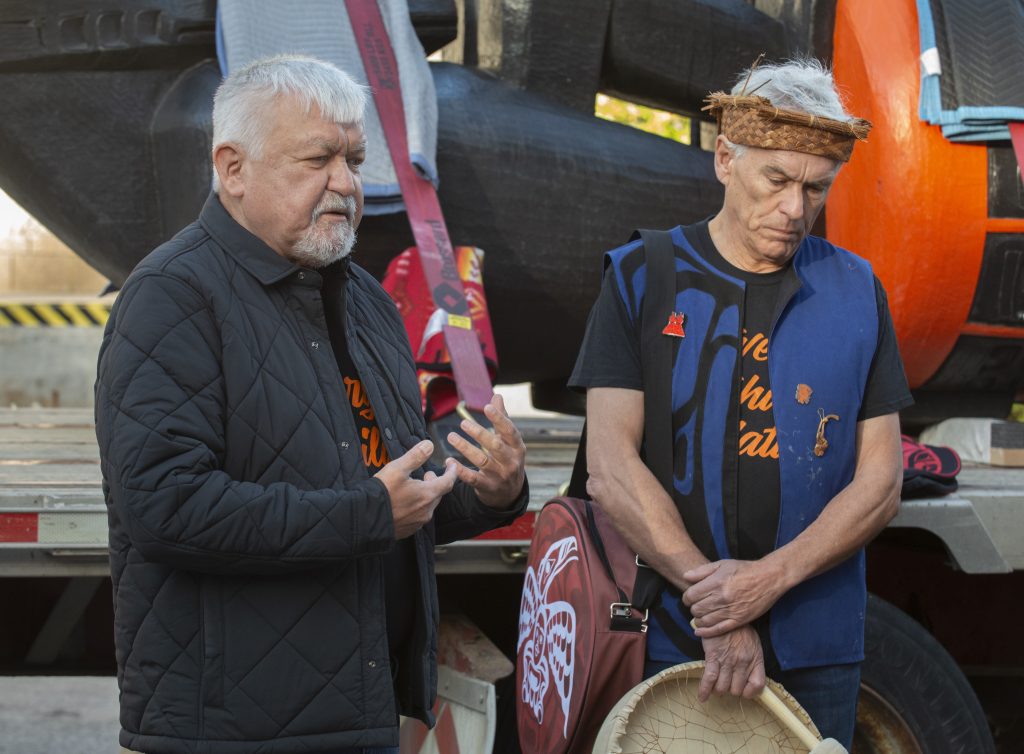 Two medium-skinned men with white hair stand in front of a large orange and black carving.