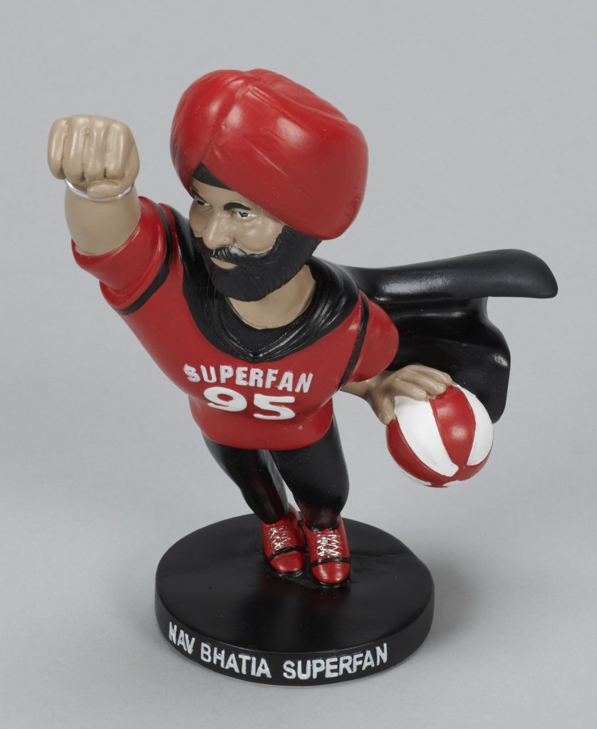 A plastic toy figure of a dark-skinned man with a black beard, wearing a red turban, a red shirt saying “Superfan 95,” and a black cape, and holding a red and white basketball.