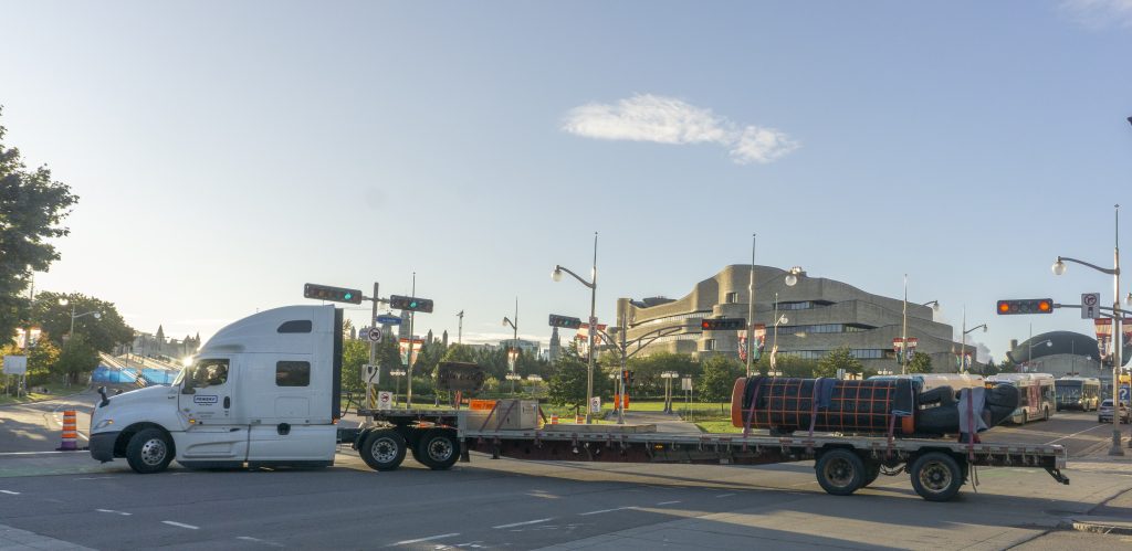 A transport truck passes in front of the Museum building, carrying the Monument lying down on a long trailer.