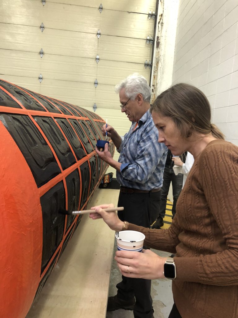 A few people stand next to a large, horizontal, cylindrical carving. They are applying paint with small brushes.