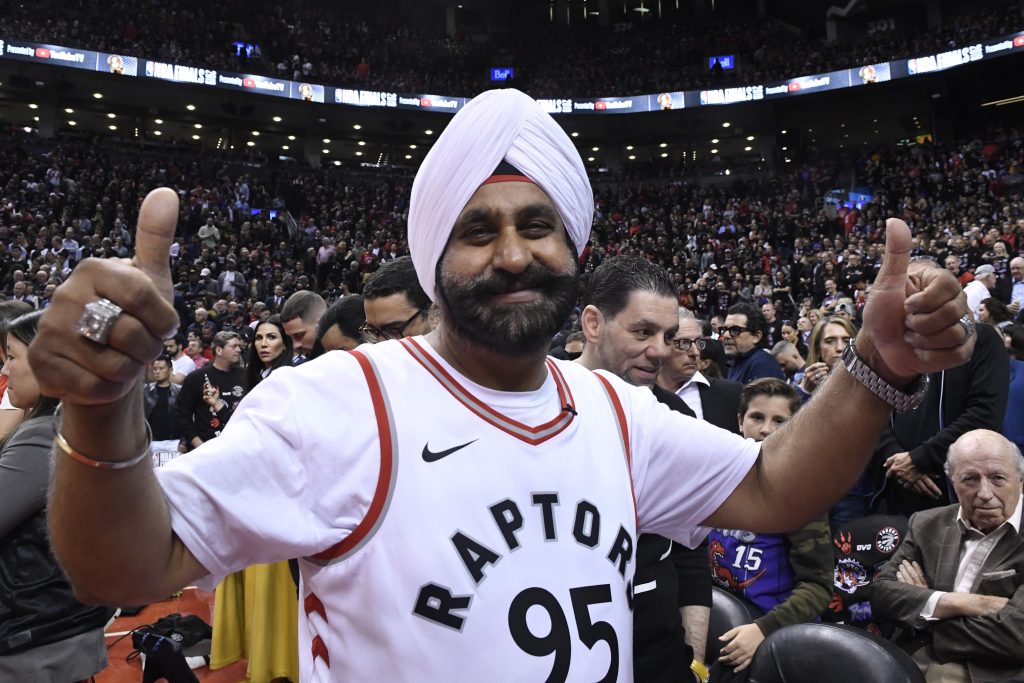 A dark-skinned man with a black beard wears a white turban and a white athletic shirt saying “Raptors 95” while giving two thumbs up and smiling at the camera. In the background is a large stadium full of people.