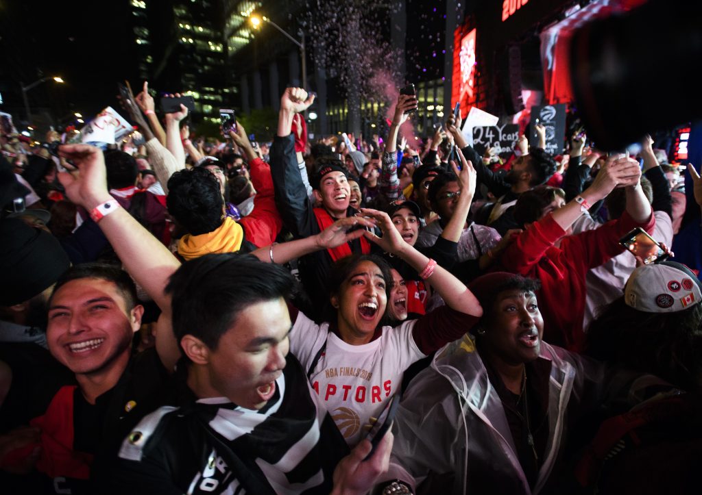 A large crowd of people gathered outside at night among large buildings cheer and raise their hands in the air.