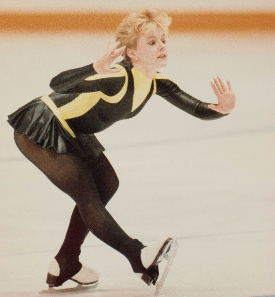 A blonde woman in a yellow and black leotard and short skirt skates while bending forward and gesturing with her hands.