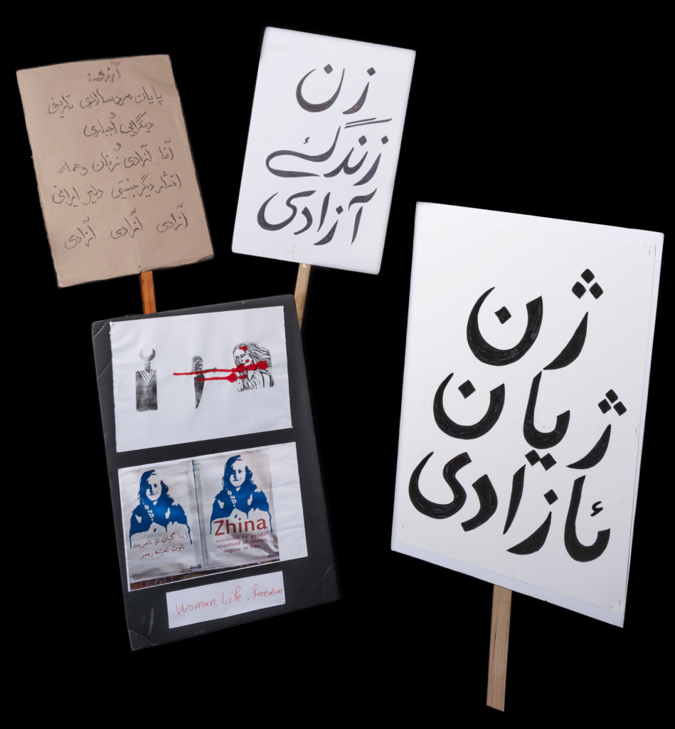 A group of placards and signs, some with writing in Kurdish and Farsi, one with a blue monochrome print of a face above the name “Zhina,” and another with a series of stylized figures connected with red paint that looks like dripping blood.