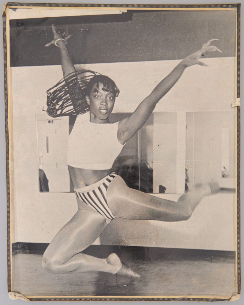 An athletic Black woman leaps into the air, arms spread wide. She has long braids that are flying to the side and wears a crop top and dance leggings.