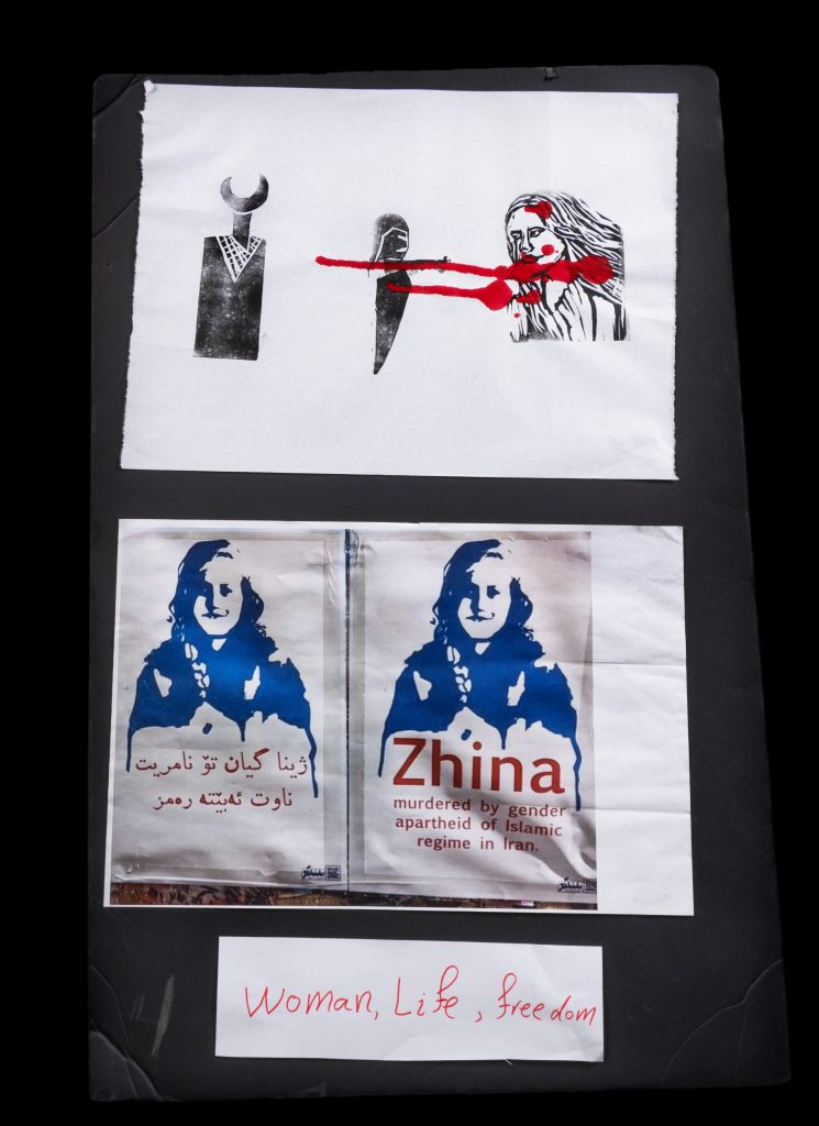 Three posters: one in block-print style, showing stylized figures connected with red paint that looks like dripping blood; one with a monochrome print of a face above the words “Zhina: murdered by gender apartheid of Islamic regime in Iran”; and a smaller handwritten sign saying “Woman, Life, Freedom.”