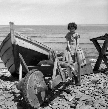 Petite fille assise sur un baril prs d'un bateau de pche sur la rive, [19--]., © MCC/CMC, J15267