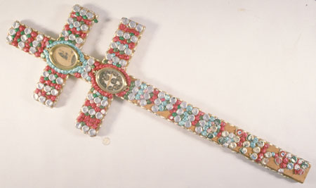 This cross is made of pine and covered in bottle caps, most of which are covered with coloured foil. There are two pictures on the cross pieces, in gold plastic frames. Some of the bottle caps are from WWII vintage beer bottles with the logo 
