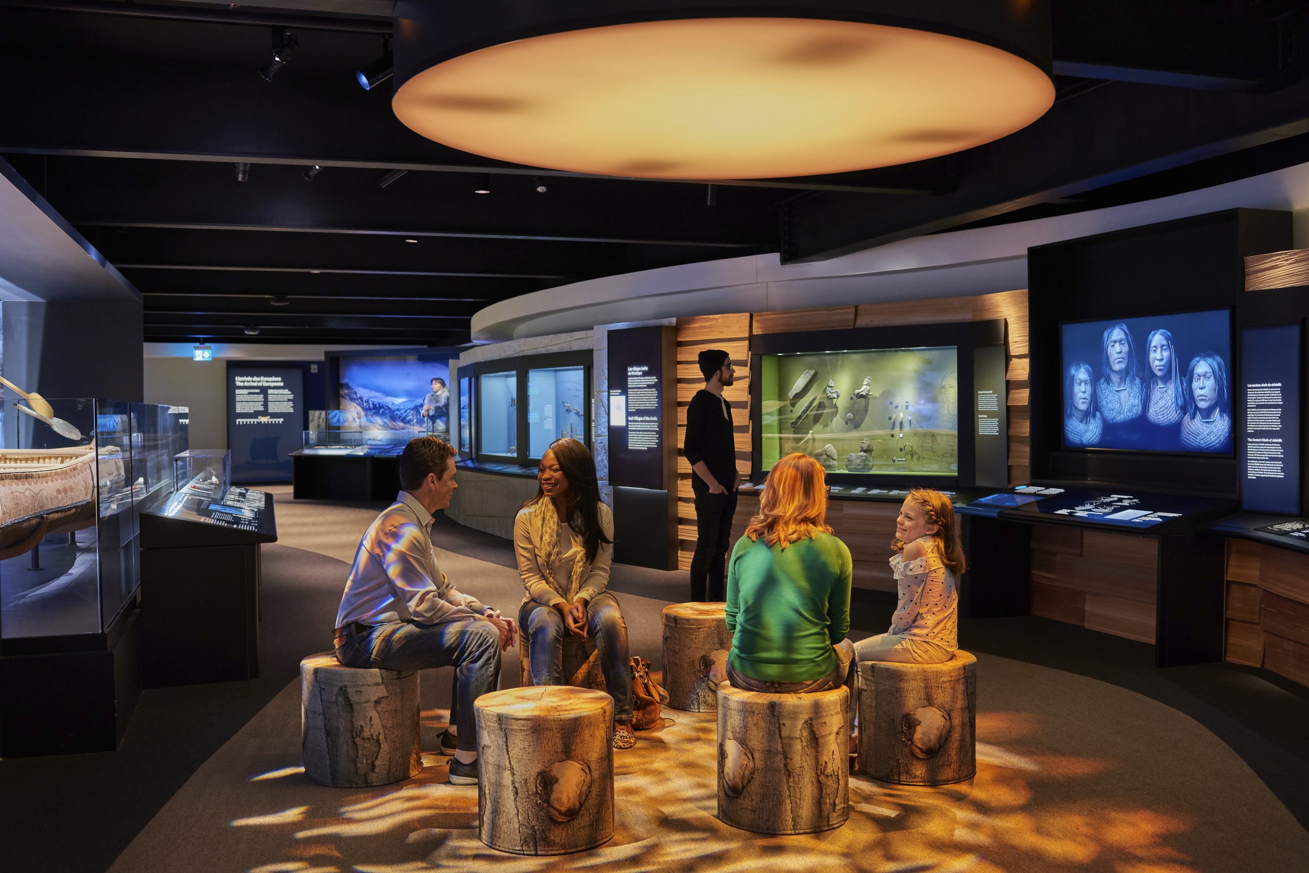 A group of people are sitting on log chairs in the Canadian Museum of History exhibit in Ottawa.