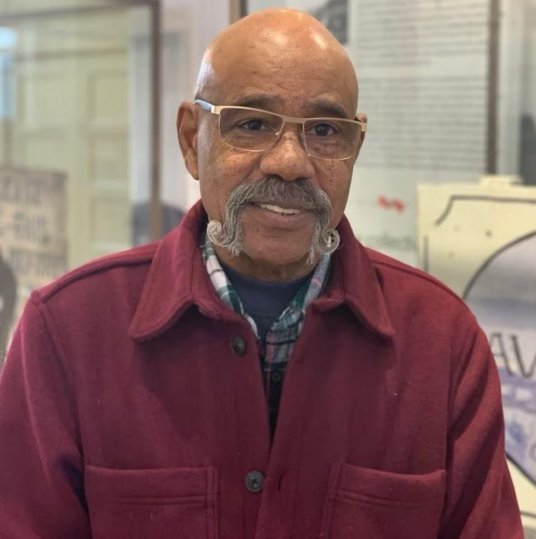 Percy Paris in a red jacket and glasses standing in front of a wall.