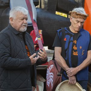 Two medium-skinned men with white hair —one holding an Indigenous drum — standing in front of a large orange and black carving.