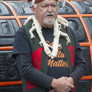 A medium-skinned man wearing leather and fur headwear, fur necklace, and cloth vest covered with simple, colourful patterns, standing outdoors in front of a large orange and back carved surface.