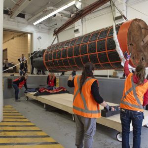 A large cylindrical sculpture is hoisted off a flatbed with wide straps. It features children’s faces painted black within an orange grid. A large carved black raven looks down upon the children from the top.