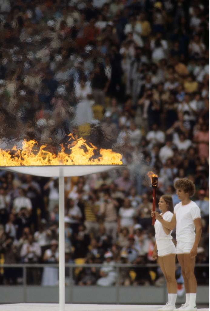 Two young people lighting a large cauldron from a torch with a large crowd in the background