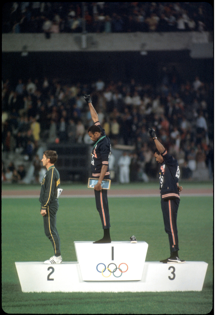 Three people on an Olympic podium, two of them with one fist up in the air, in a stadium with a large crowd in the background.