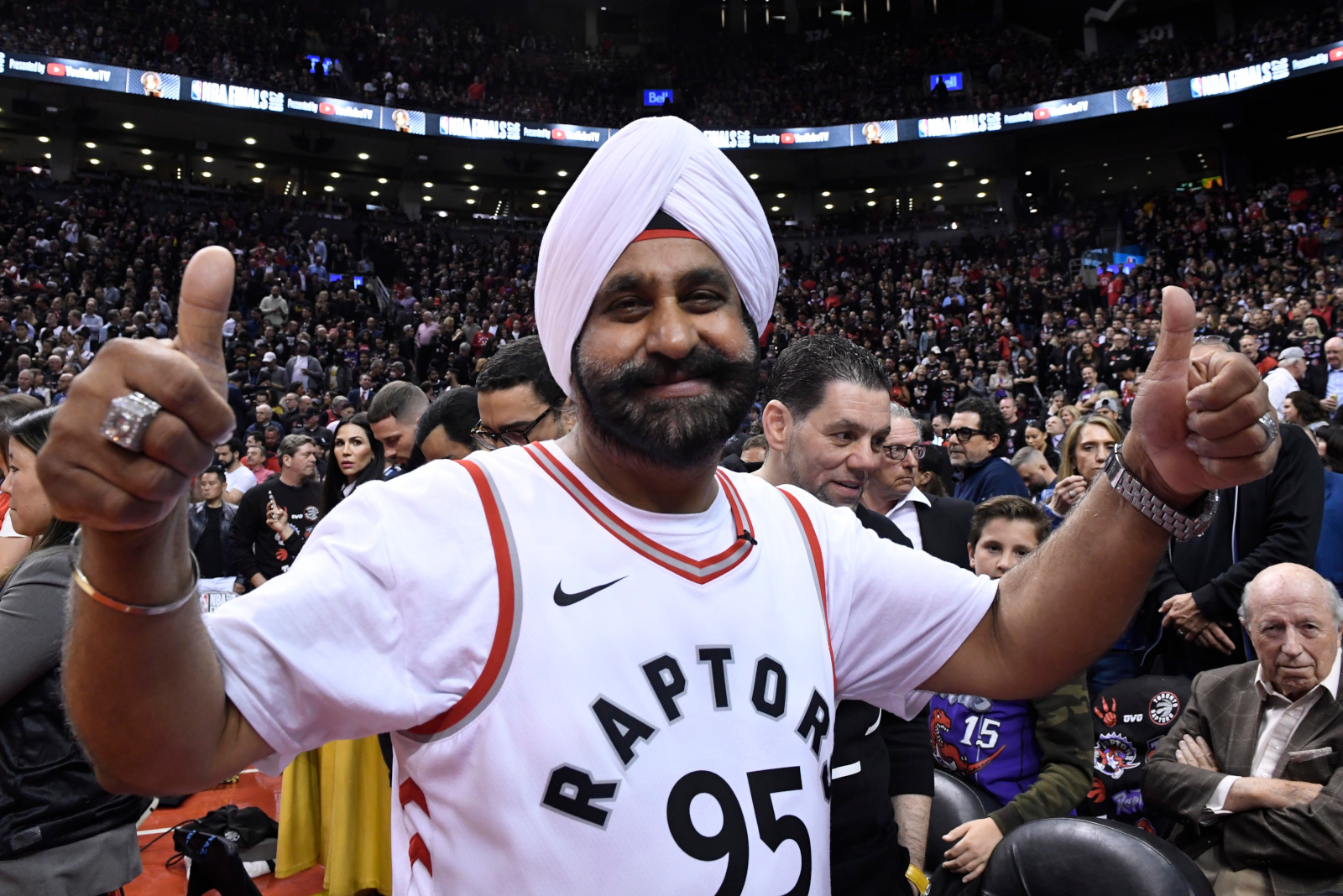 A dark-skinned man with a black beard wears a white turban and a white athletic shirt saying “Raptors 95” while giving two thumbs up and smiling at the camera. In the background is a large stadium full of people.