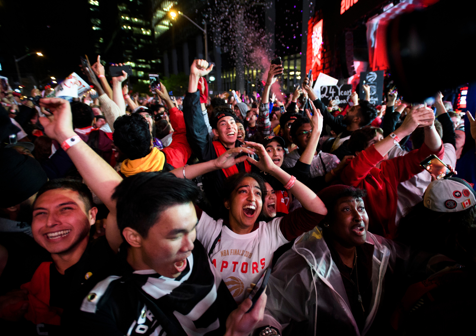 A large crowd of people gathered outside at night among large buildings cheer and raise their hands in the air.