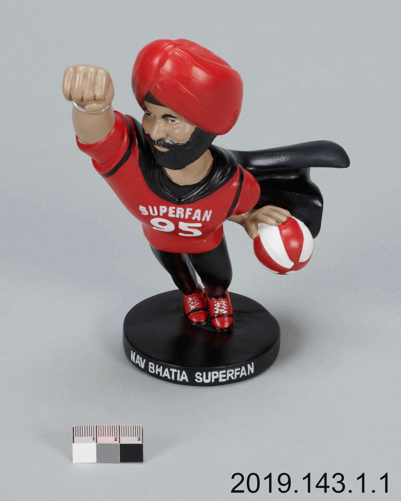 A plastic toy figure of a dark-skinned man with a black beard, wearing a red turban, a red shirt saying “Superfan 95,” and a black cape, and holding a red and white basketball.