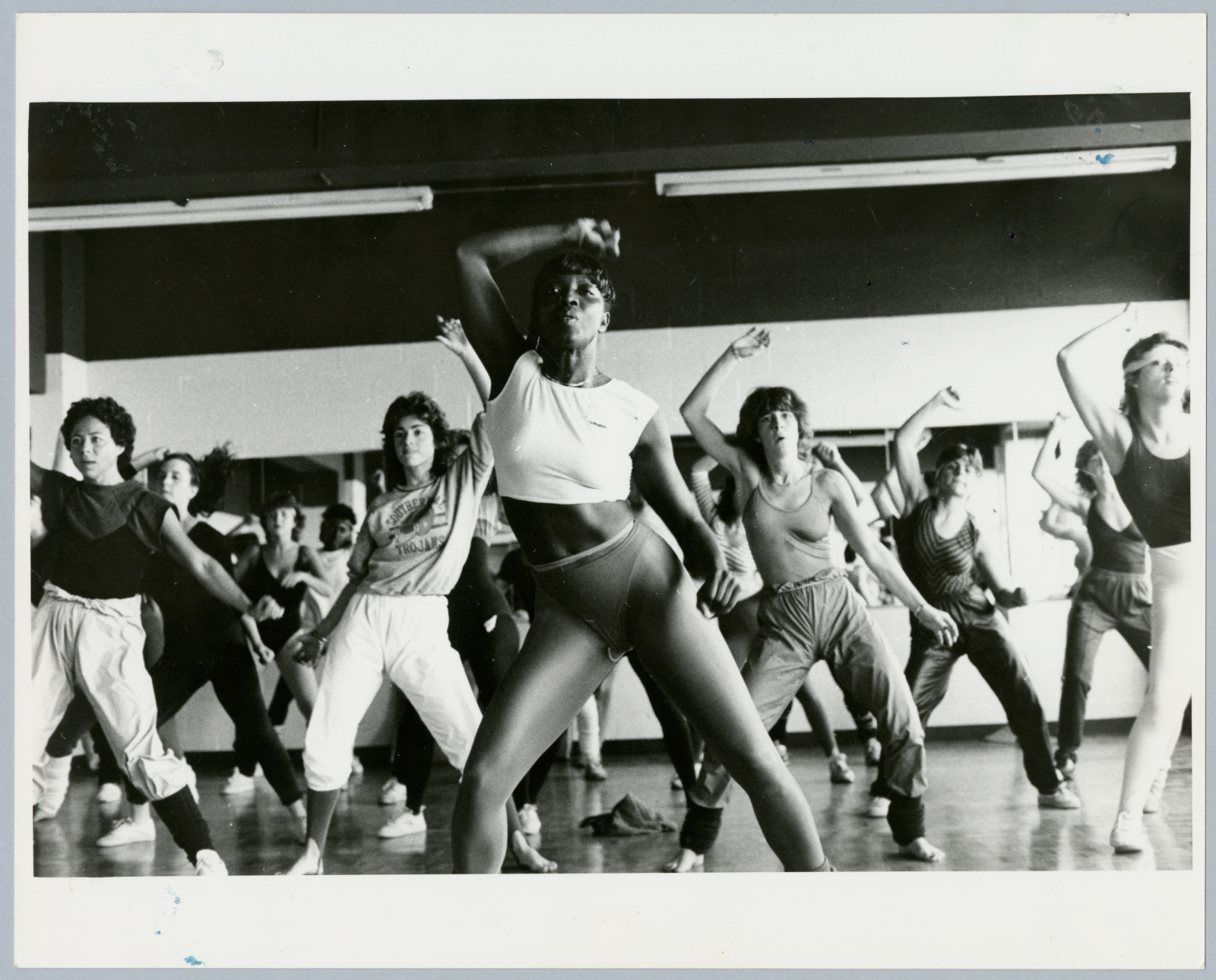 A crowded fitness class with people wearing a variety of fitness clothes, led by an athletic Black woman in a crop top and leggings.