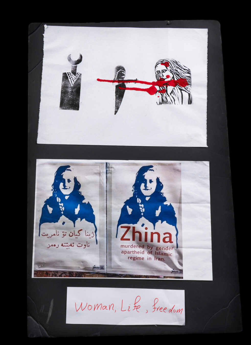 Three posters: one in block-print style, showing stylized figures and connected with red paint that looks like dripping blood; one with a monochrome print of a face above the name “Zhina”; and a smaller handwritten sign saying “Woman, Life, Freedom.”