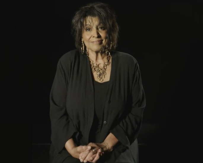 A smiling Black woman wearing a black top, black cardigan, a gold-coloured necklace, and hoop earrings.