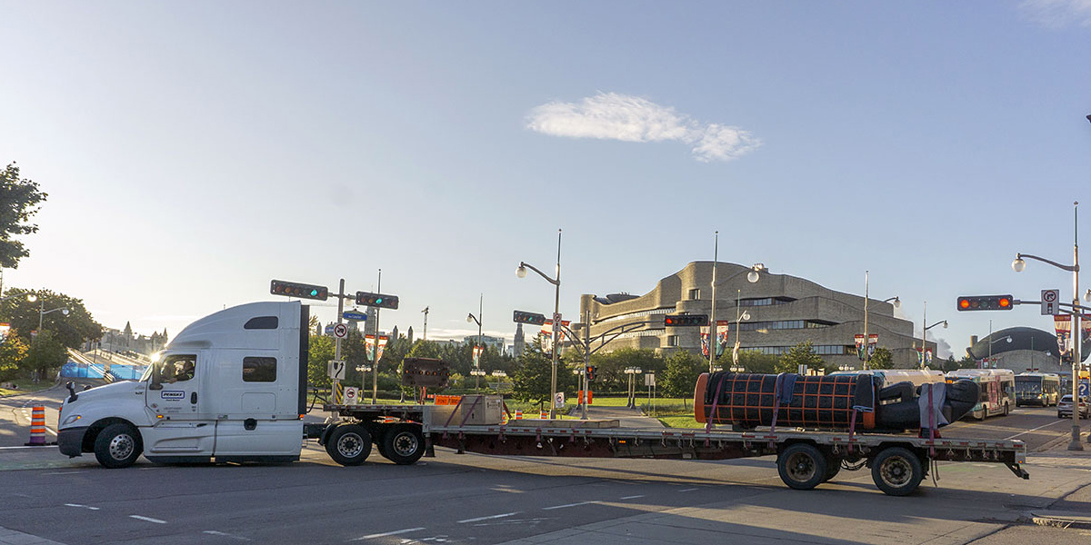 Large truck in front of a sandstone-coloured Museum building, with the Monument lying on a long trailer.