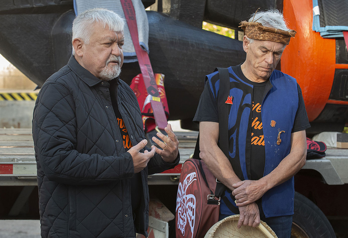 Two medium-skinned men with white hair —one holding an Indigenous drum — standing in front of a large orange and black carving.