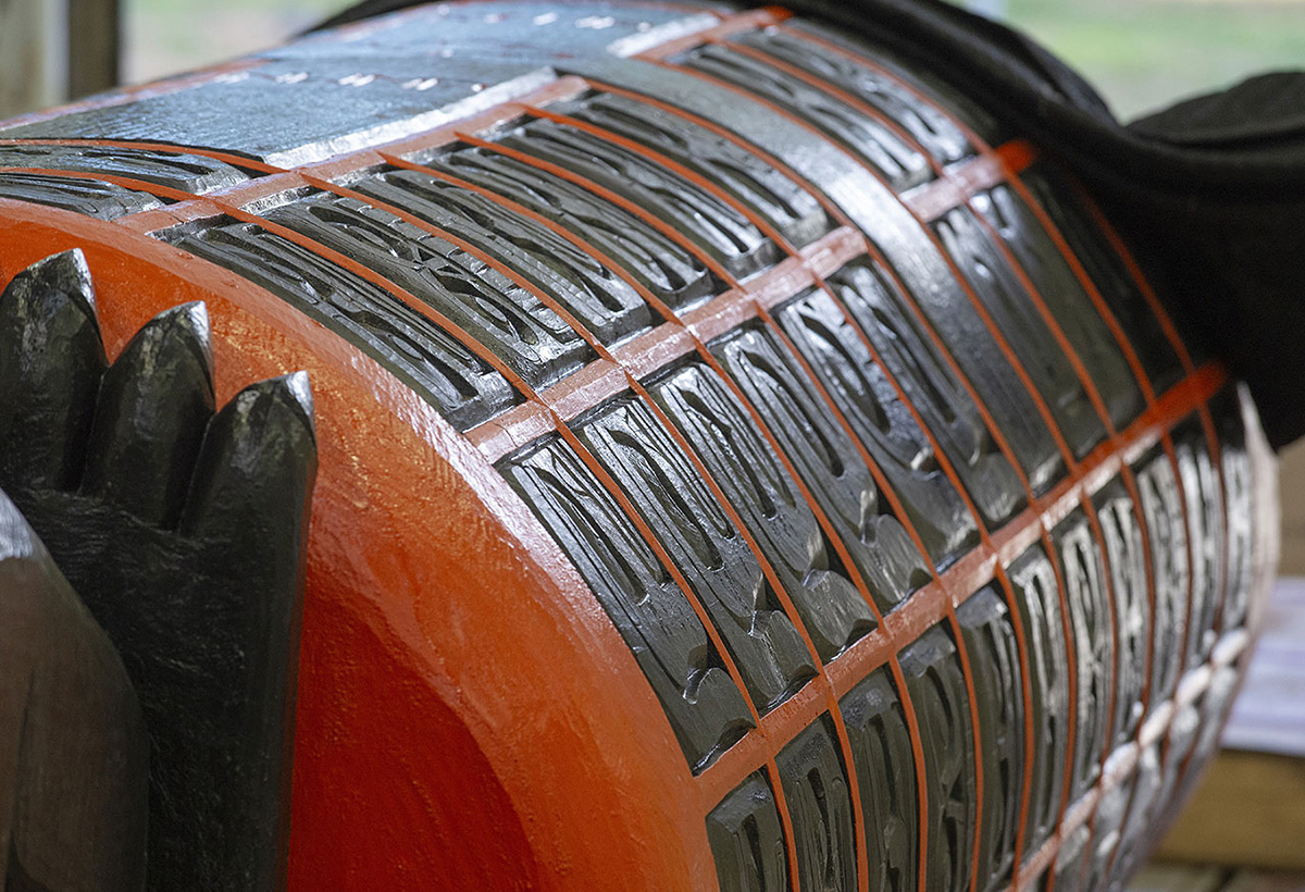 A view of the lower body of the Monument, lying on its side. The large cylindrical shape is covered with carved faces set within a bright orange grid.