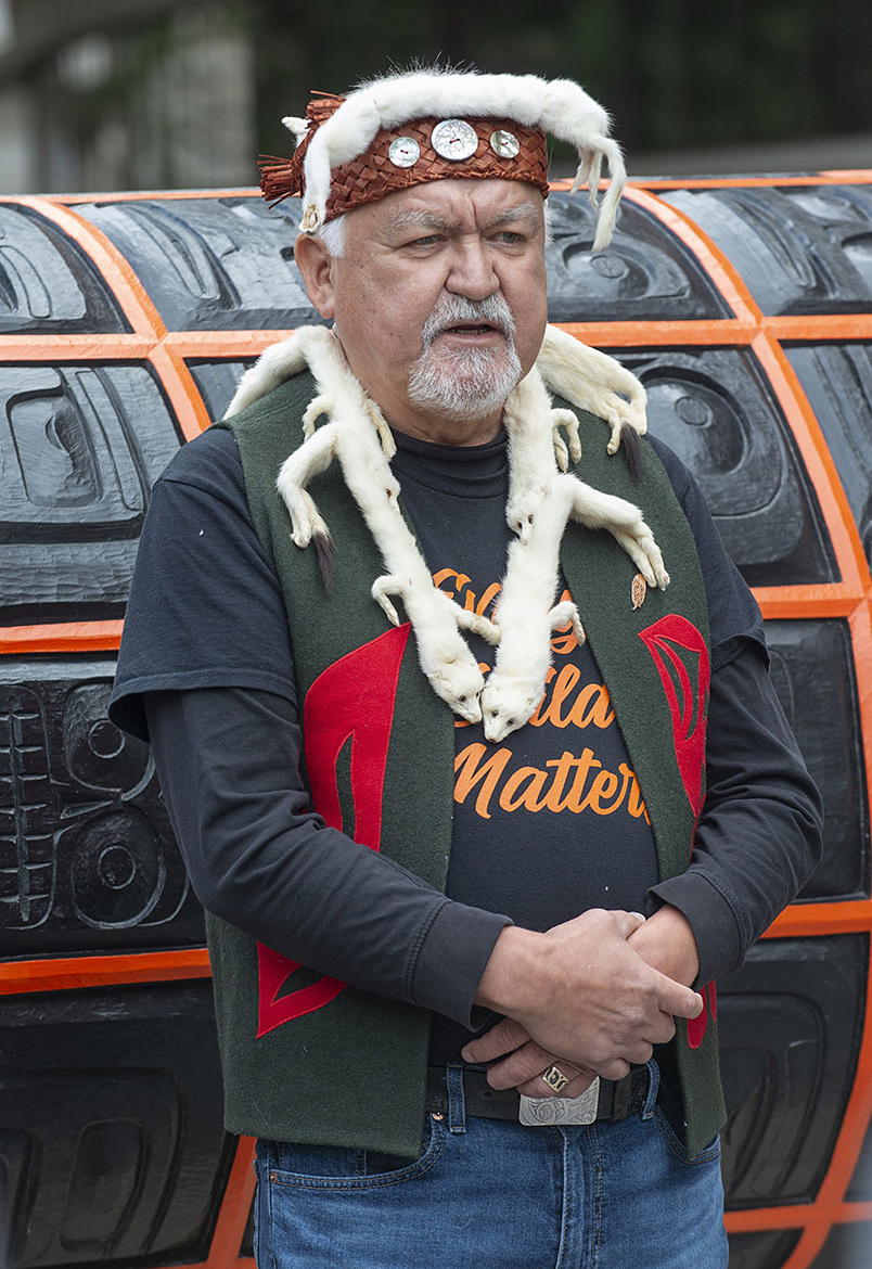 A medium-skinned man wearing leather and fur headwear, fur necklace, and cloth vest covered with simple, colourful patterns, standing outdoors in front of a large orange and back carved surface.