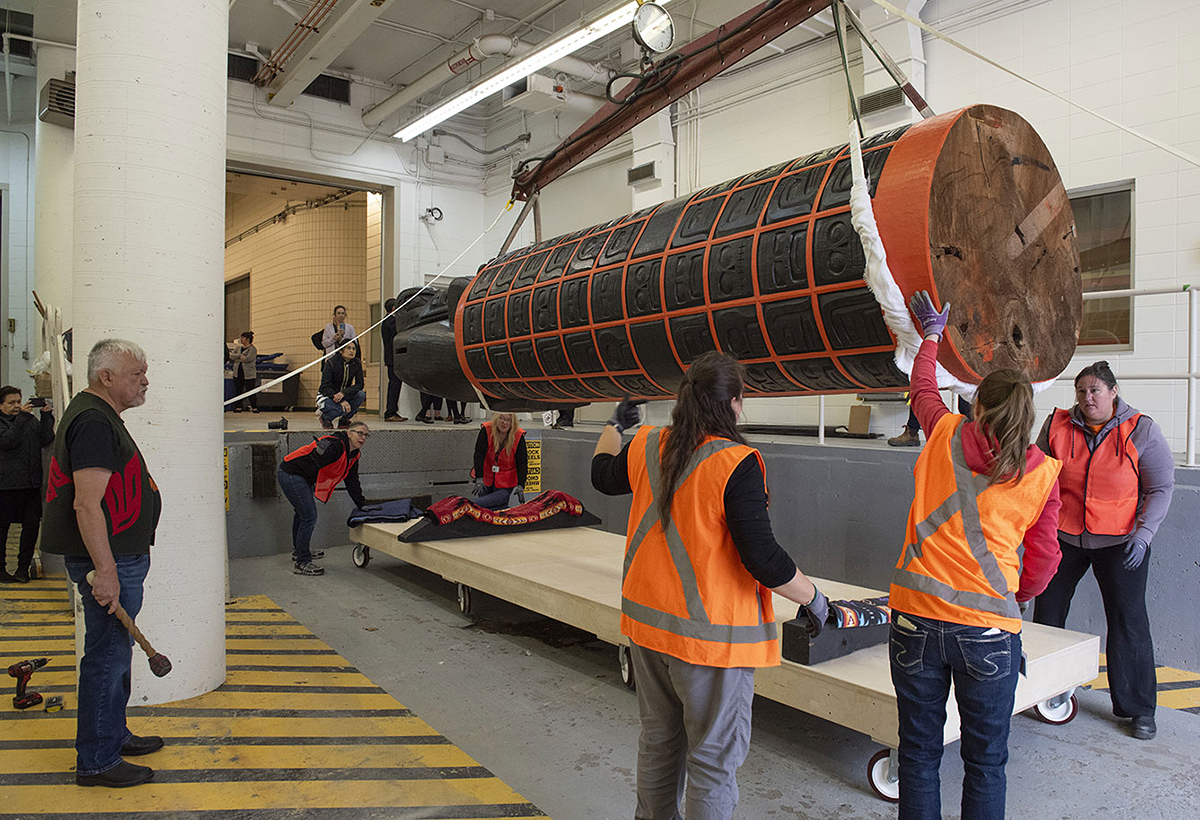 A massive carved and painted wood sculpture being hoisted onto a platform inside a loading dock. Workers in bright orange vests help, while others look on.