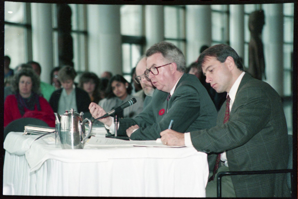 Photograph of two men in suits seated at a table signing a document, with people seated in the background