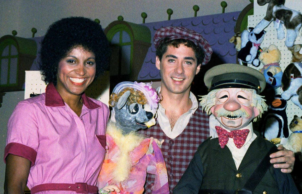 A candid photo of a Black woman wearing a pink jumpsuit; a small puppet of a grey and white animal wearing colourful clothes; a light-skinned man wearing a plaid vest and flat cap; and a life-sized puppet of a light-skinned older man in a bow tie, cap and jacket.