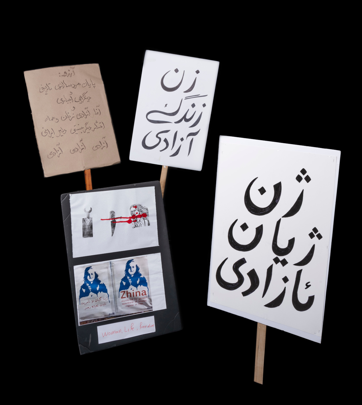 A group of placards and signs, some with writing in Kurdish and Farsi, one with a blue monochrome print of a face above the name “Zhina,” and another with a series of stylized figures and connected with red paint that looks like dripping blood.