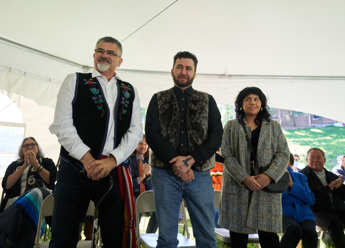 Three people stand side by side in a white festival tent outside. From left to right, a light-skinned man with a salt-and-pepper goatee and very short hair wears a beaded vest and a colourful dangling sash; a light-skinned person with dark-brown beard and hair wears a grey vest and jeans; a shorter medium-skinned person with long hair wears a grey plaid long coat and a shoulder bag.