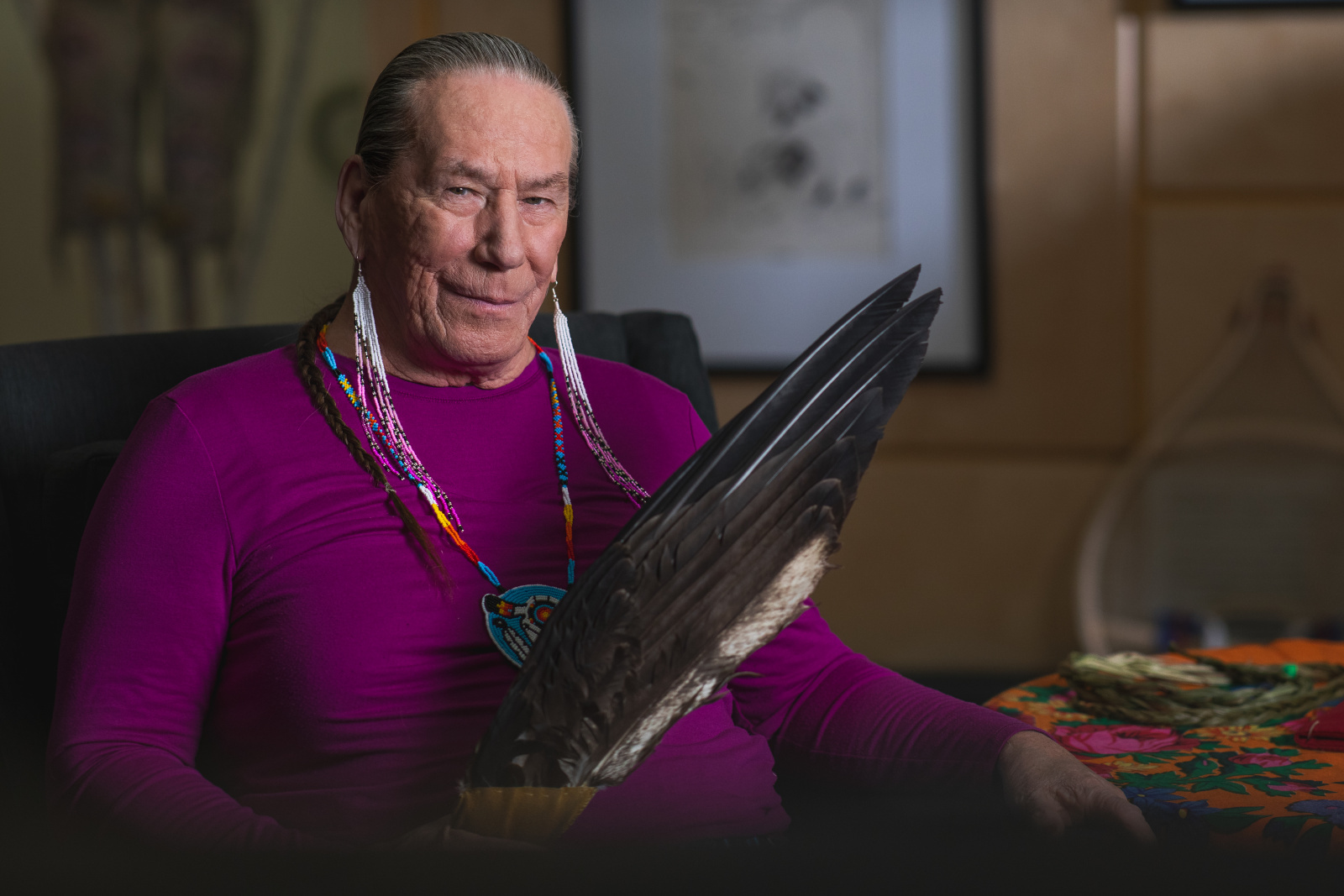 A person wearing a purple top and long beaded necklaces sits in a chair, holding an eagle feather fan.