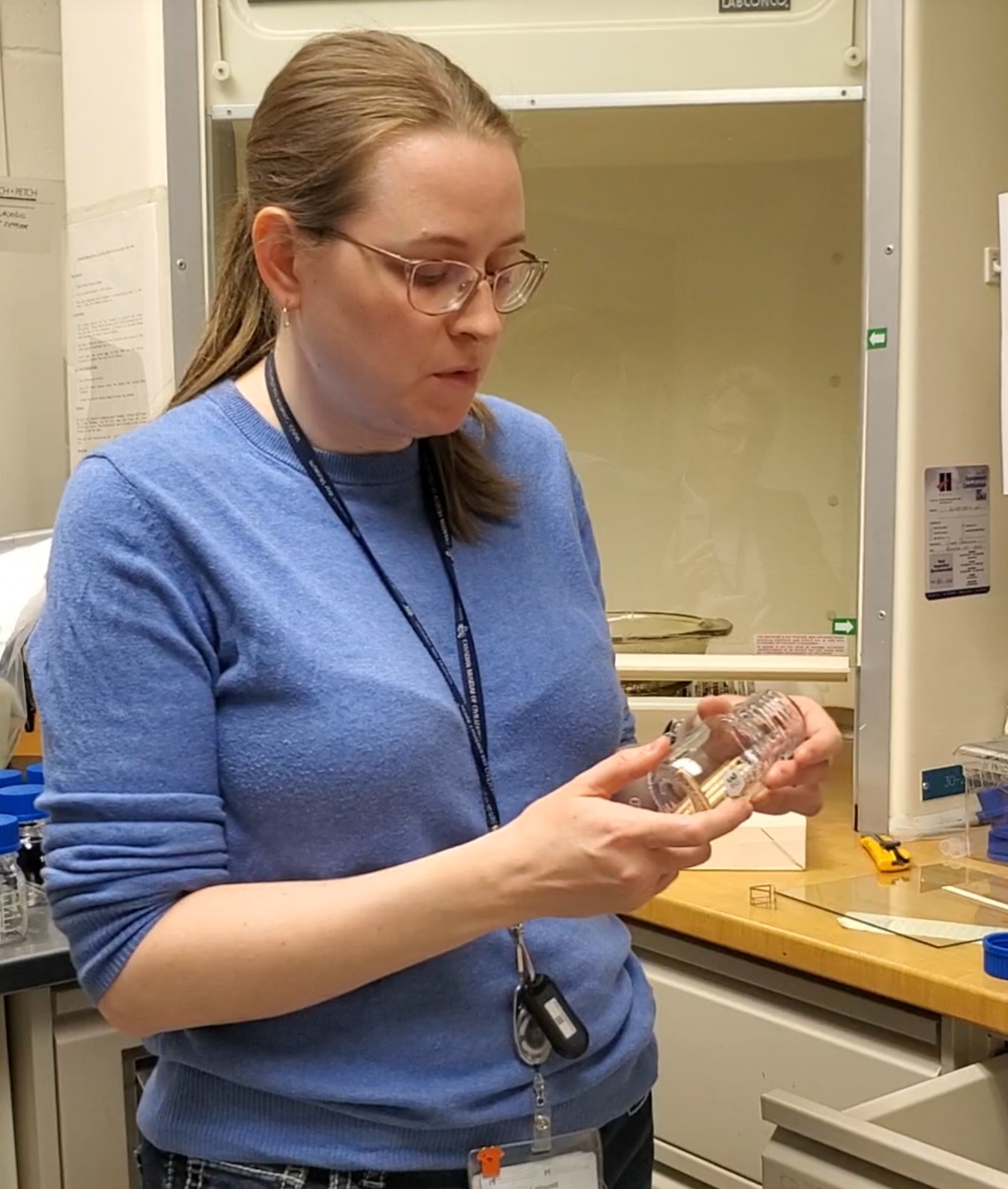 A person with a long ponytail stands in front of laboratory equipment, holding a small glass jar.