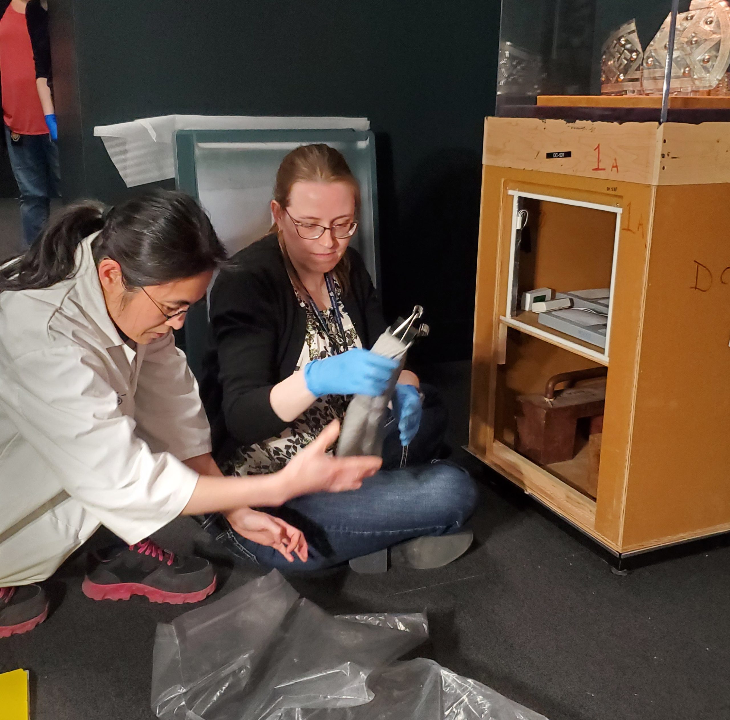 Two people kneel in front of a display case with its sides removed, showing some equipment inside.