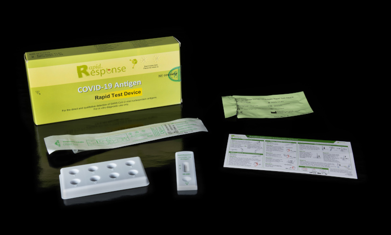 A small cardboard box reading “Rapid Response COVID-19 Antigen Rapid Test Device” next to its contents: a flat, rectangular piece of plastic with eight round holes, a small rectangular test unit, two foil-wrapped items, and a small card of instructions.