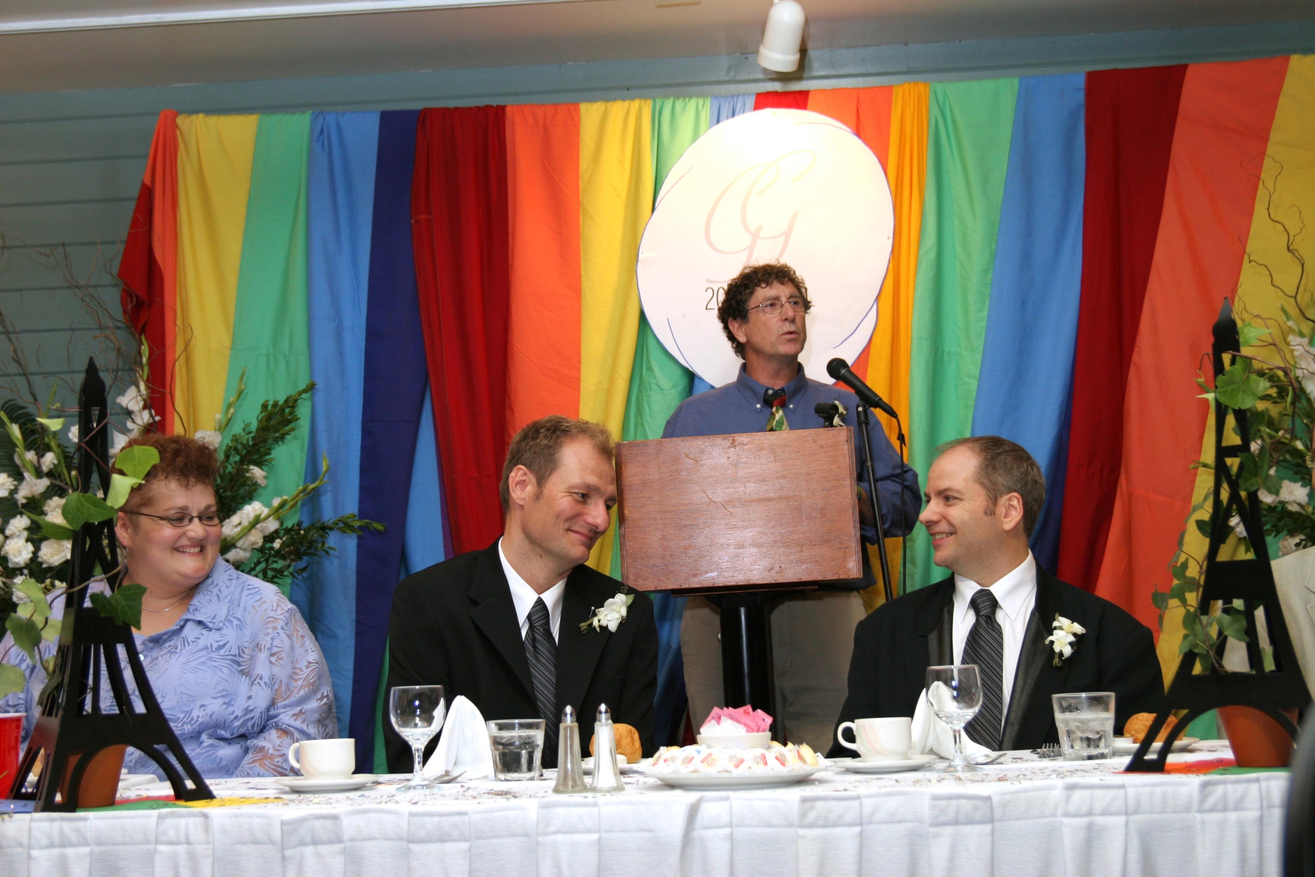 Two men sit at a head table, a man speaks at a podium behind them in front of a rainbow backdrop
