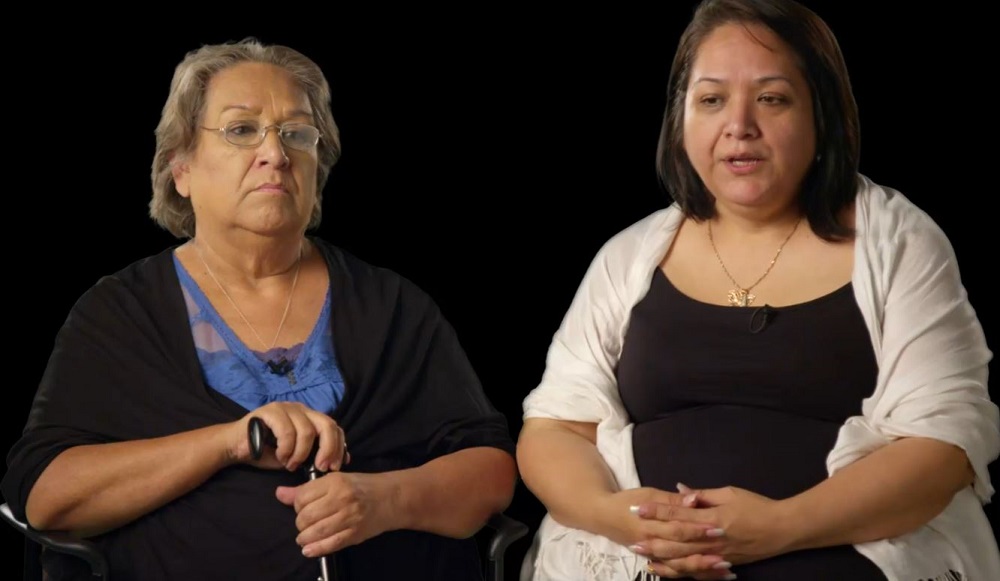 A still from a video, showing two women seated next to each other. One woman is an older adult with a medium skin tone and medium-length grey hair. She is wearing glasses and a blue shirt with a black cardigan, and is holding a walking stick. The woman next to her is an adult with shoulder-length dark hair and a medium skin tone. She is wearing a black shirt with a white sweater and a necklace.