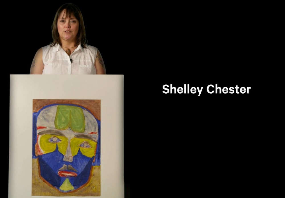 A still from a video, showing an adult woman with medium skin tone and shoulder-length dark hair. She is standing and holding a large piece of art. The art features a multicoloured mask.